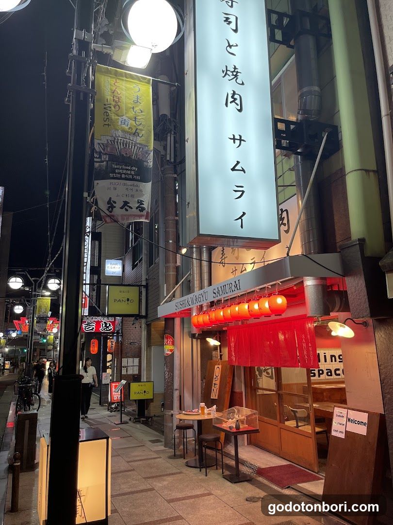 Night view of a Japanese restaurant storefront with vertical signage reading 'SUSHI YAKITORI SAMURAI', red lanterns, and outdoor seating area.