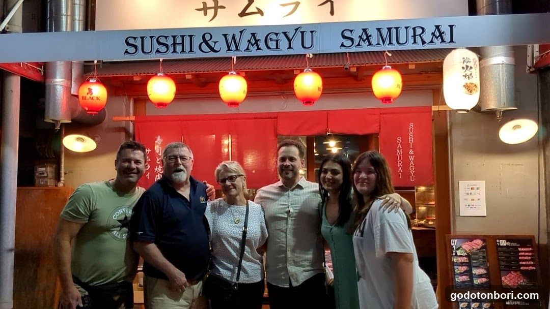 A group of six people stands in front of a Japanese-style restaurant entrance. The storefront features a prominent sign reading 'SUSHI & WAGYU SAMURAI' with hanging lanterns and red curtains.