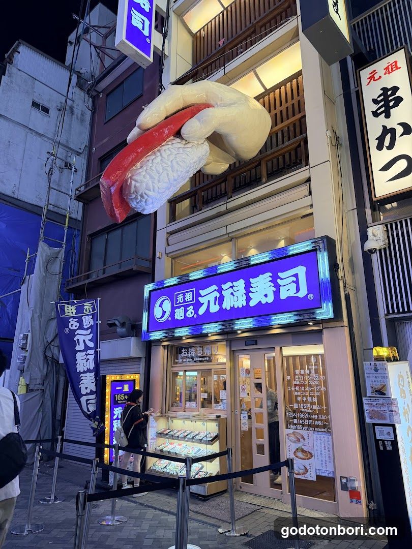 Night view of a sushi restaurant storefront with illuminated signage reading '元禄寿司'. A large sculpture of a hand holding sushi hangs above the entrance. Glass display cases show sushi items inside, and menus are posted on the door.