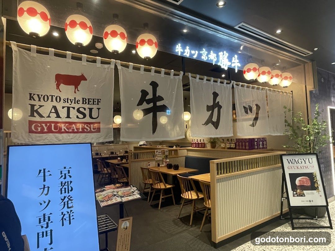 A Japanese restaurant storefront featuring hanging white noren curtains with bold black kanji characters and a red cow logo. Above, glowing paper lanterns line the entrance. A blue illuminated sign displays vertical Japanese text. To the right, a freestanding menu board advertishes Wagyu Gyukatsu.