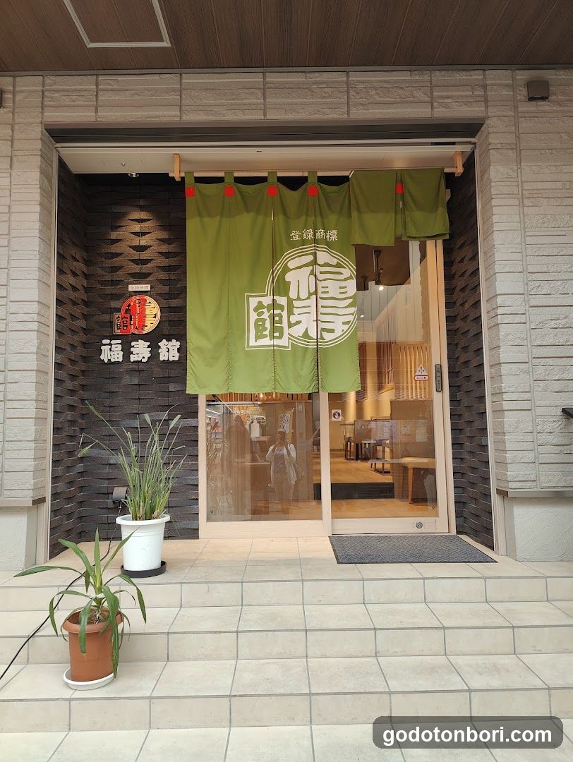 The image shows the exterior entrance of a shop named 'Fukusho no Tei' (福寿館). The facade features dark brick walls with white lettering spelling out the name. A green noren curtain hangs above the glass doors, displaying Japanese characters and the same name in large stylized font. Two potted plants are placed on the tiled steps leading to the entrance. Through the glass door, the interior appears warm and wooden, suggesting a traditional or casual dining atmosphere.