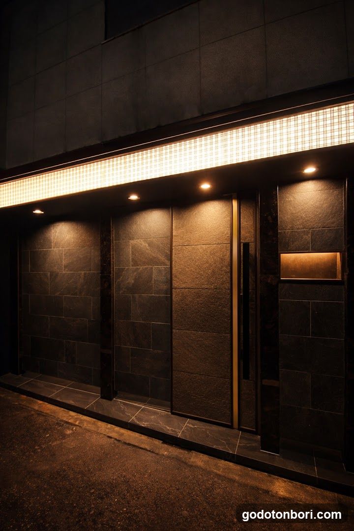 A modern storefront entrance featuring dark stone cladding and recessed lighting. A horizontal illuminated grid sign runs above the door, casting a warm glow on the textured walls. The entrance includes glass doors with vertical handles and a small rectangular plaque mounted to the right wall.