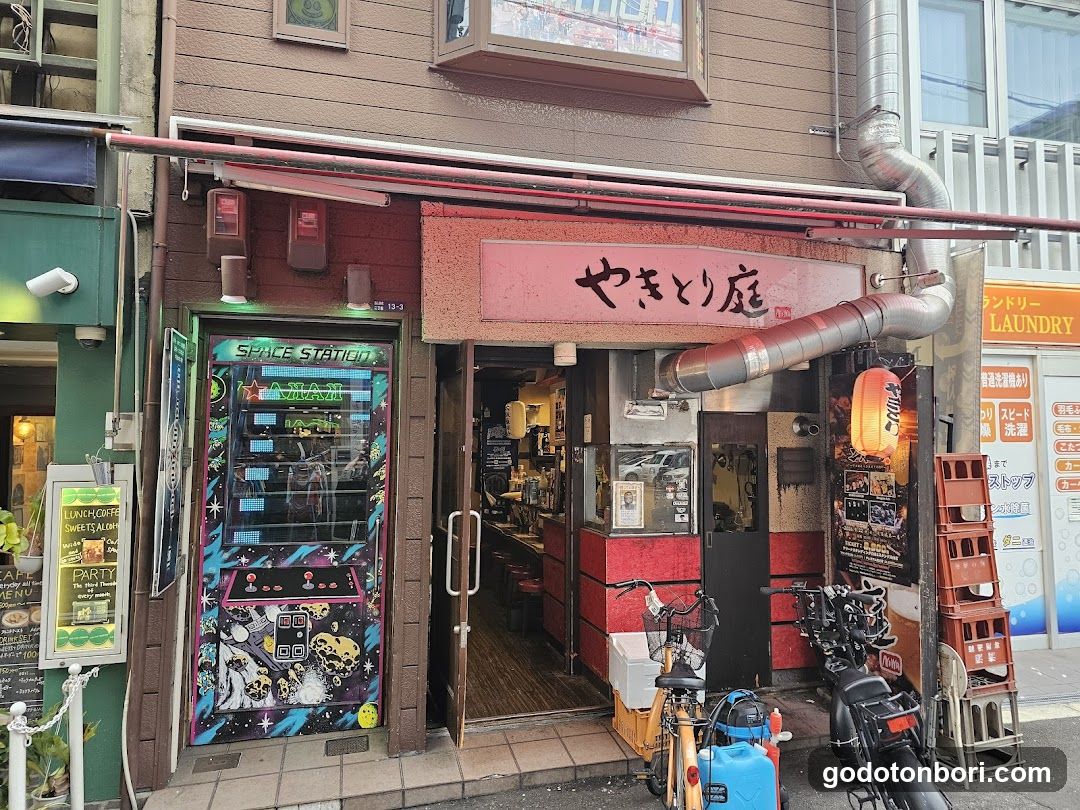 A street-level view of a Japanese yakitori restaurant named 'Yakitori Tei' (やまとり庭). The facade features a prominent red sign with black calligraphy above the entrance. To the left is an arcade game cabinet labeled 'SPACE STATION'. A large silver ventilation duct runs vertically along the right side. In front, there are stacked orange crates and parked bicycles/motorcycles. An adjacent building to the right has laundry service signage.