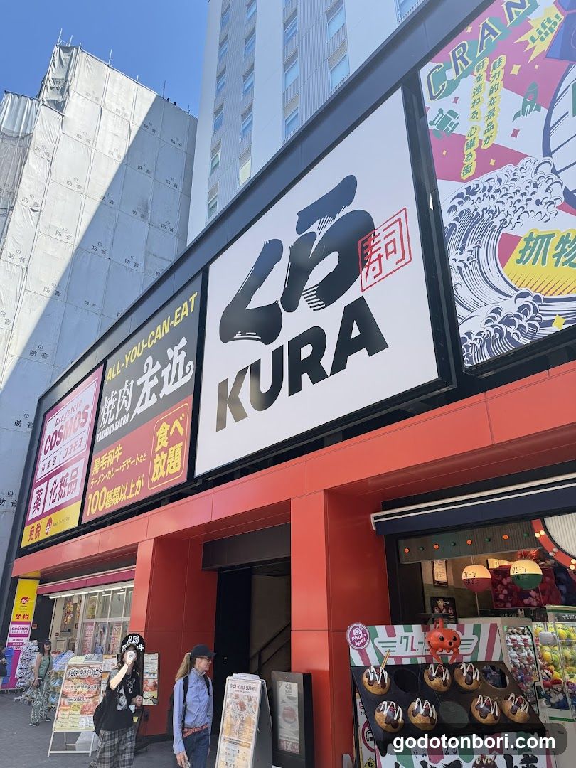 A street-level view of a restaurant named 'KURA' with prominent signage featuring Japanese characters and English text. The building facade is red, with multiple signs advertising food items like grilled meat and sushi. A display stand outside showcases small food items on sticks.