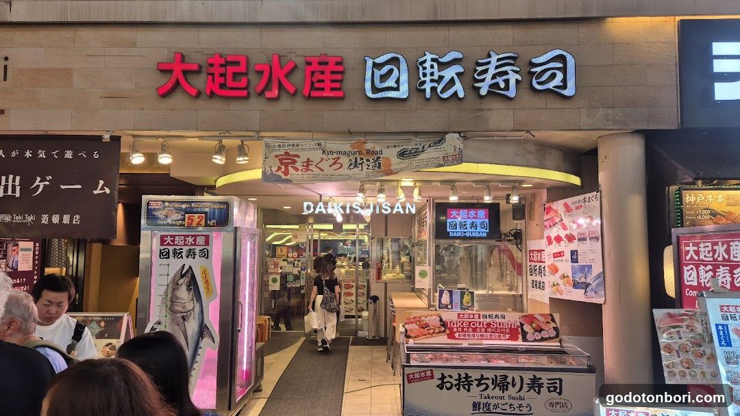 A brightly lit storefront of a Japanese回转寿司 (rotational sushi) restaurant, featuring large red and white signage above the entrance. The facade includes illuminated signs in both Japanese characters and English ('DAIKISUISAN'). A vertical display case on the left showcases a whole fish with promotional graphics. To the right, there are additional posters advertising sushi rolls and promotions like 'Takeout Sushi'. People are visible entering or standing near the entrance.