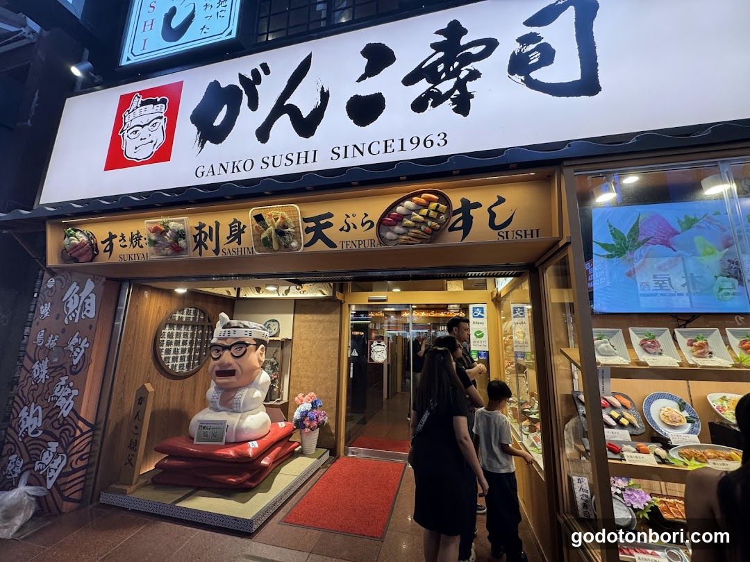 A brightly lit Japanese sushi shop with a large white sign reading 'GANKO SUSHI SINCE1963' and stylized kanji characters. Below, a wooden panel displays images of sushi rolls and tempura with Japanese text labels like 'SUSHI', 'TENPURA', and 'SUKUYA'. A life-sized statue of the chef character stands on red cushions near the entrance. The glass door is open, revealing interior lighting and customers inside. To the right, a display case shows various sushi items under warm lighting.