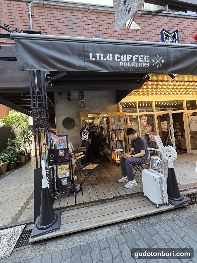 A coffee shop with a black awning displaying 'LILO COFFEE ROASTERS' in white text. The storefront features wooden flooring, outdoor seating, and a person sitting on the porch. There are various signs and posters near the entrance.