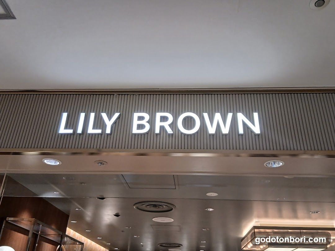 The image shows the illuminated sign of a store named 'LILY BROWN' mounted on a modern, metallic facade with vertical slats. The lighting is bright and evenly distributed, highlighting the brand name against the neutral background.