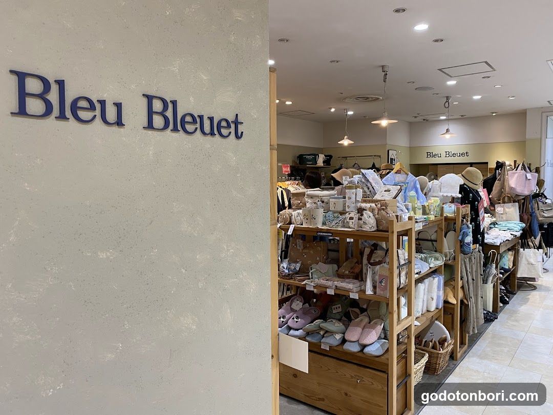 Interior view of a retail store with wooden shelving displaying folded clothing, slippers, and accessories. The left wall features the brand name 'Bleu Bleuet' in blue lettering. A second sign reading 'Bleu Bleuet' is visible on the back wall.