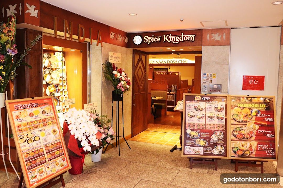 Entrance to Spice Kingdom restaurant with visible signage, floral arrangements, and menu boards displaying food items.