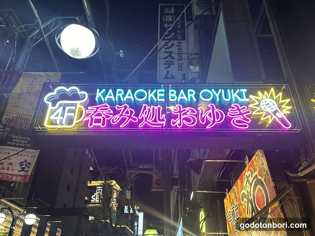 Night photo of a brightly lit neon sign above the entrance to Karaoke Bar Oyuki, featuring Japanese and English text with microphone and beer mug graphics.
