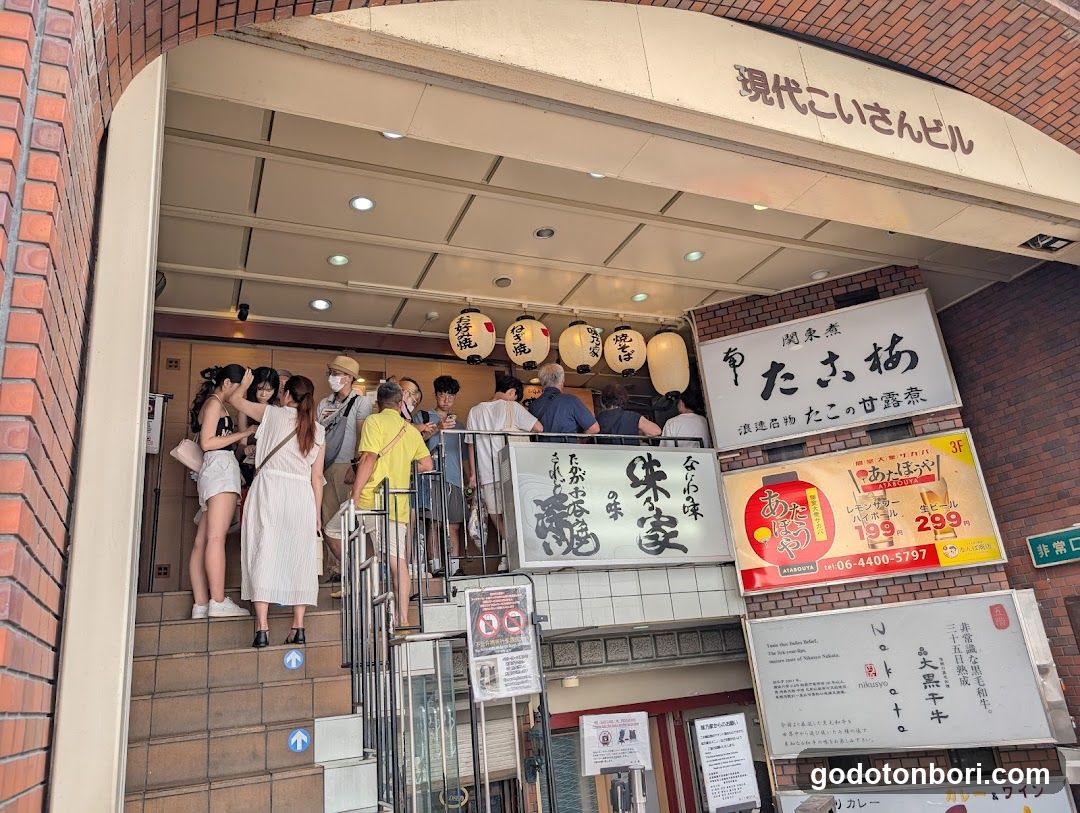 A busy entrance to a Japanese building with multiple signs, including 'Kawashima Tsubaki' and 'Takeshiya'. People are gathered near the entrance, some ascending stairs. Lanterns hang above, adding traditional decor.