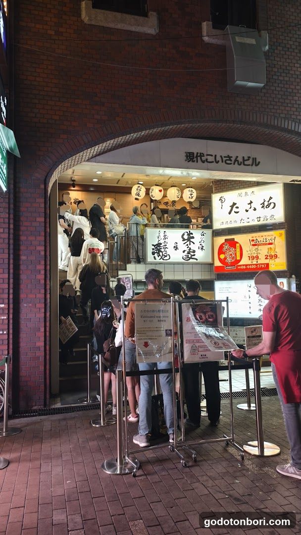 A brick building entrance with a large arched doorway leading to an indoor dining area. The sign above reads '現代こいさんビル' (Modern Kisan Building). Inside, customers are seated at tables under paper lanterns. Outside, people queue behind metal stanchions near a menu board displaying prices like ¥198 and ¥239.