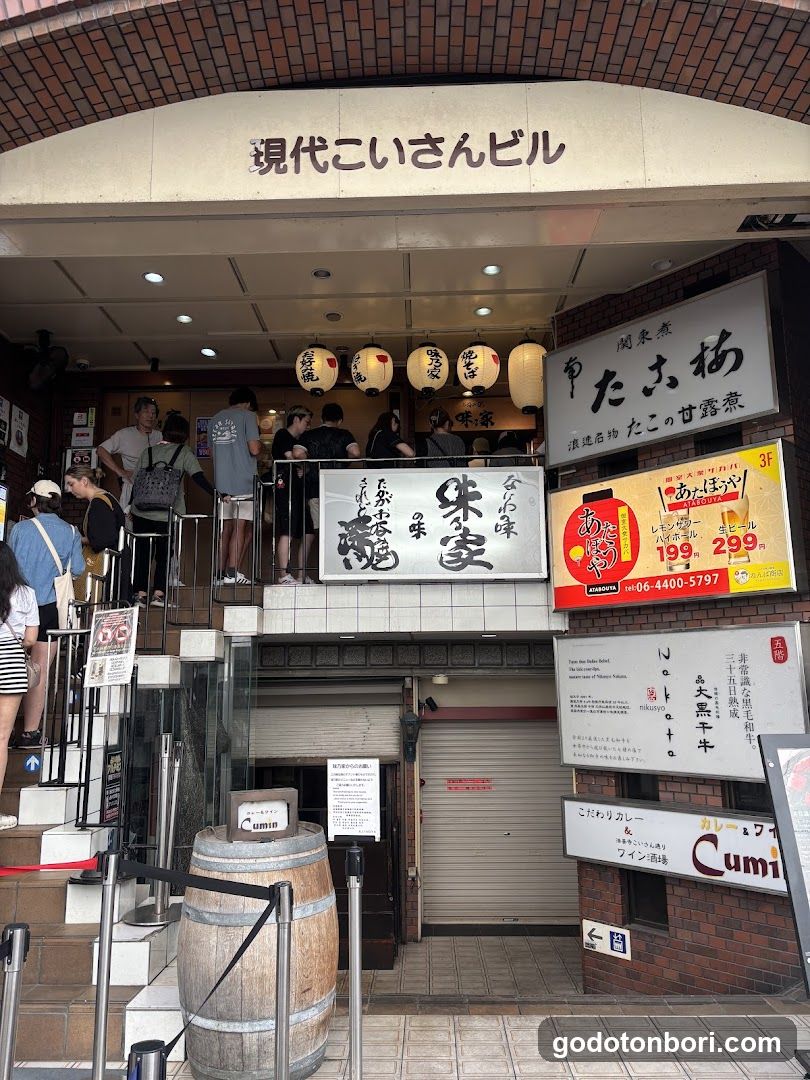 The image shows the exterior entrance of a building with multiple signs and banners. The main sign reads '現代こいさんビル' (Kodai Koisan Building). Below, there are several smaller signs including one that says 'たのしみ味 朱家' (Tanoshimi Aji Shuka) and another advertising 'あまほぼろ' (Ama Hoboro) with prices listed as 199 yen and 299 yen. There is also a barrel in front of the entrance and people standing on the steps leading up to the building.