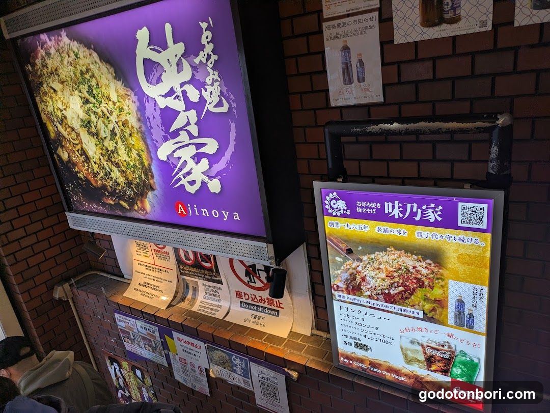 Photo of the exterior entrance showing a large illuminated sign with Japanese characters and 'Ajinoya' in English, alongside a vertical menu board featuring food images and pricing details on a brick wall.