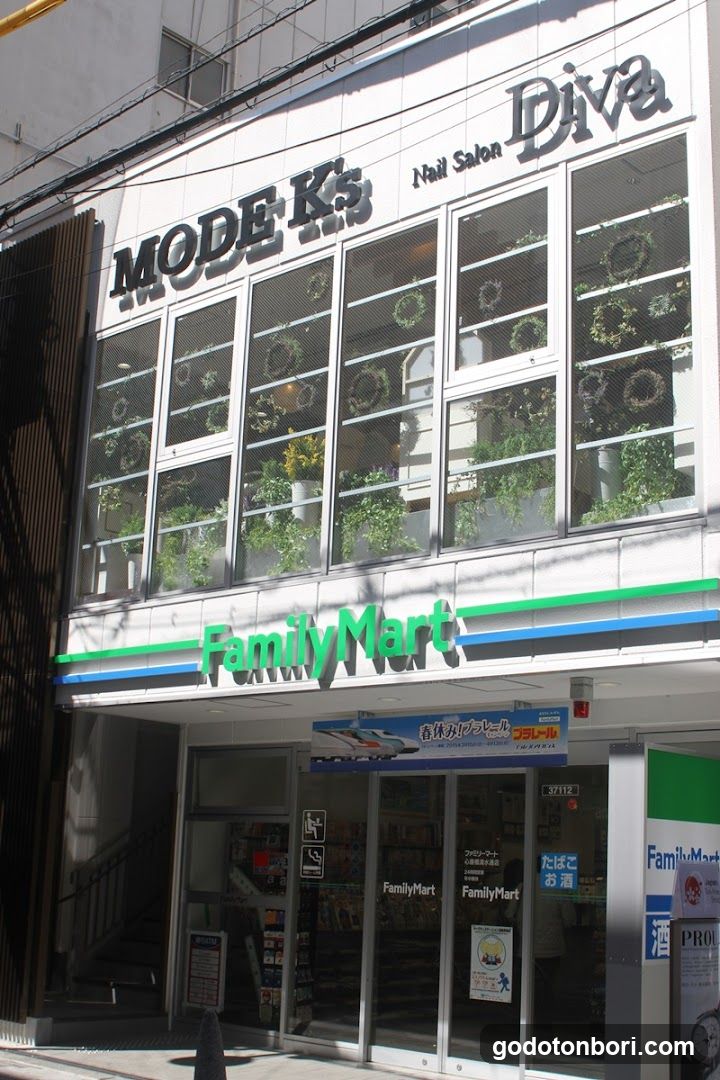 The image shows the exterior of a FamilyMart convenience store with an adjacent nail salon named 'Diva' above it. The building features large windows displaying potted plants, and signage includes 'MODE K's', 'Nail Salon Diva', and 'FamilyMart'. A blue banner advertises a train service.