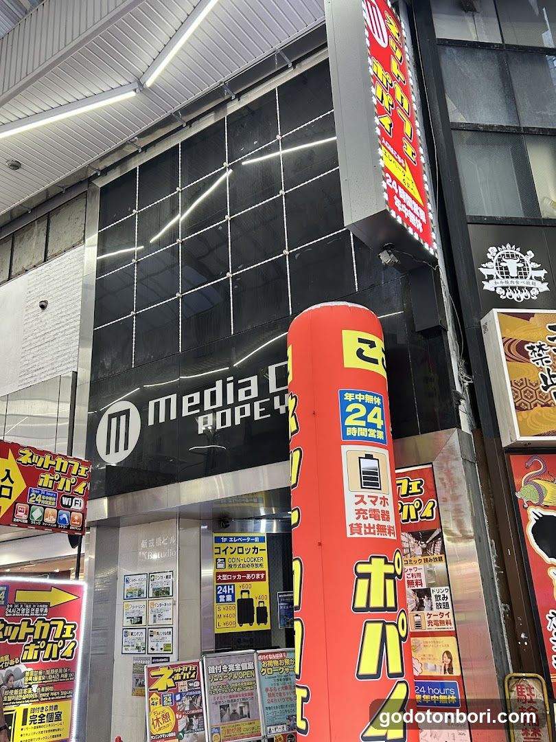 Exterior view of a shop with 'media' and 'ROPE' signage on the building facade. A large red inflatable pillar displays Japanese text including '24 hours' and 'free phone charger'. Multiple posters and advertisements are visible around the entrance, including one mentioning 'net cafe' and another advertising '24-hour open' services.