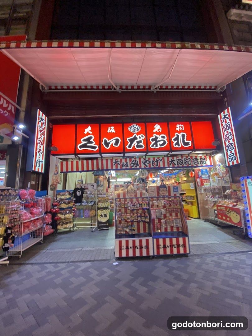 A brightly lit storefront with a red and white striped awning. The main sign displays Japanese characters in bold white on a red background, flanked by vertical banners. Outside displays feature packaged snacks and sweets arranged on shelves and racks.
