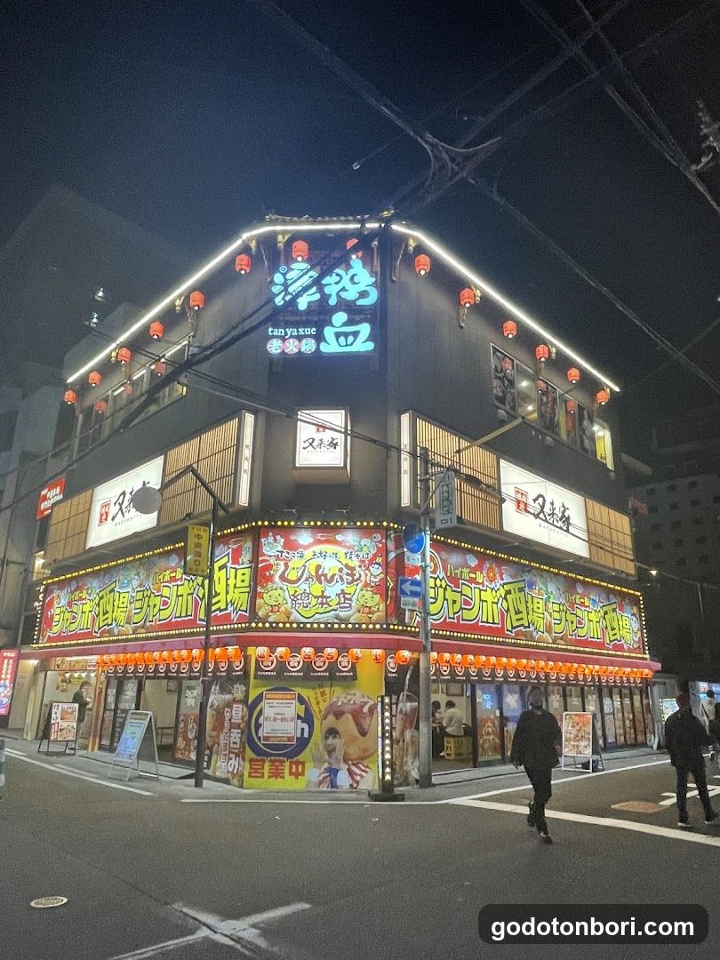 A brightly lit multi-story building at night, featuring prominent signage in Japanese characters and English text like 'tan yaku'. The ground floor has colorful banners with neon lights and red lanterns hanging along the roofline. Several people are walking on the sidewalk near the entrance.