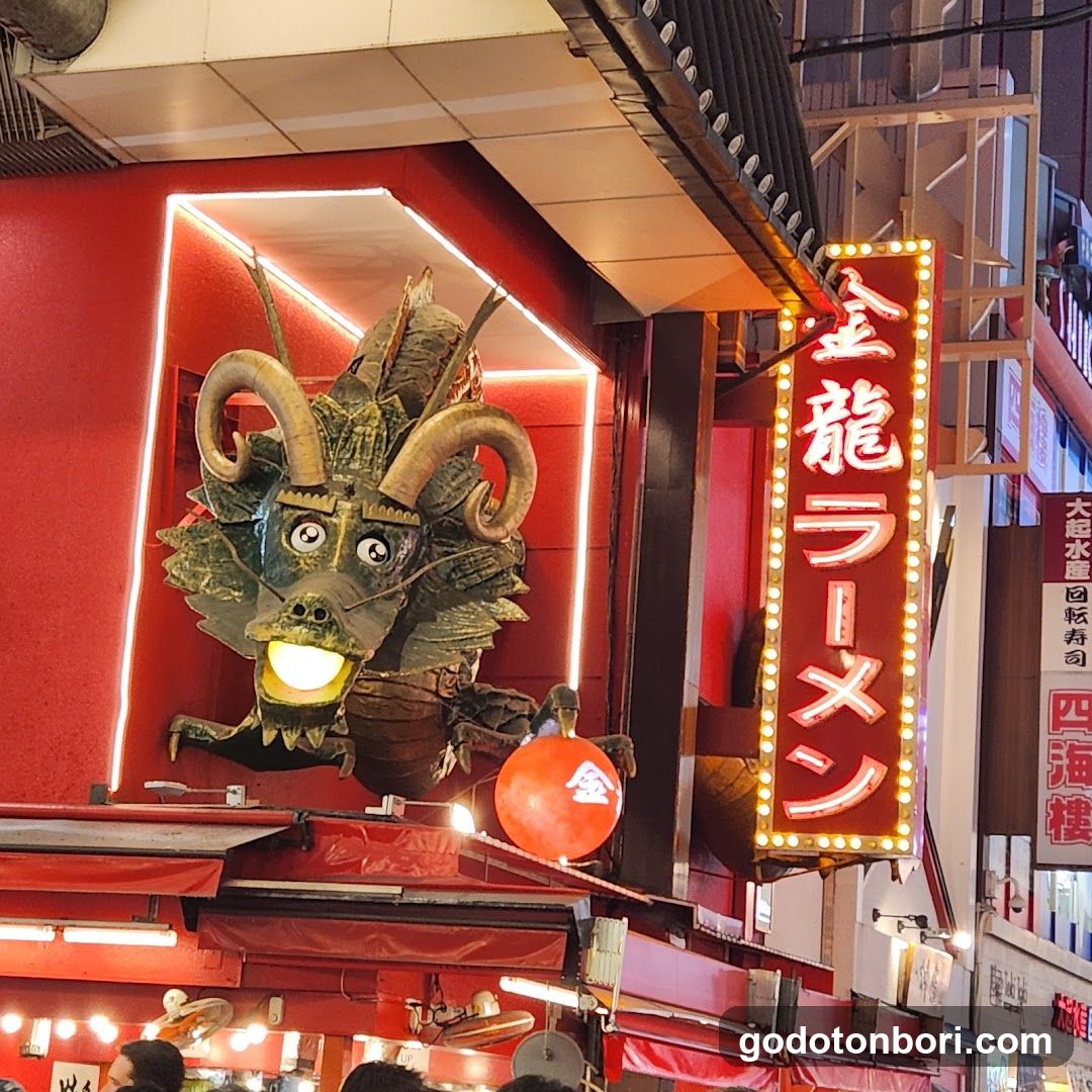 A vibrant storefront featuring a large, illuminated dragon head sculpture with glowing eyes and mouth. The building has a red facade with neon lighting outlining the dragon. A vertical sign reads '金龍ラーメン' (Kintaro Ramen) in bright lights. Below, there are additional signs and lanterns, including one with Japanese characters. The scene is lively, suggesting a bustling ramen shop.
