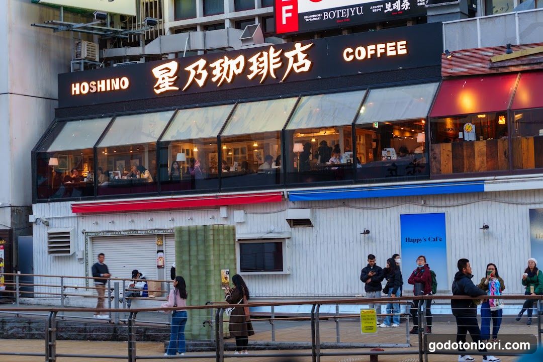 A two-story coffee shop with large glass windows on the upper level, displaying interior seating and customers. The facade features illuminated signage reading 'HOSHINO' in English and Japanese characters alongside 'COFFEE'. Below is a white building section with a closed shutter door, a small window, and people standing nearby on a walkway.