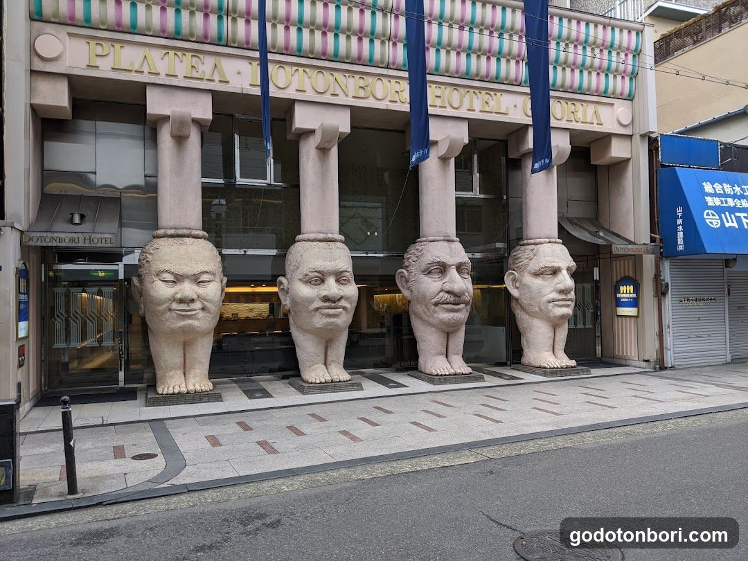 The image shows the exterior of Platea Otonbori Hotel, featuring four large stone head sculptures at its entrance. The building has a modern facade with glass windows and doors, and blue banners hanging above the entrance. A sign on the right side indicates it is part of the 'Yamaguchi Prefecture' area.