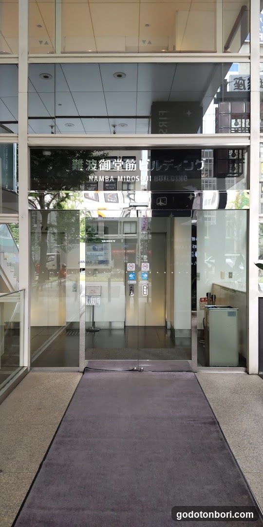 Glass entrance with signage reading '難波御堂筋ビルディング' and 'NAMBA MIDOSUJI BUILDING'. Interior visible through glass doors, including a reception desk area.