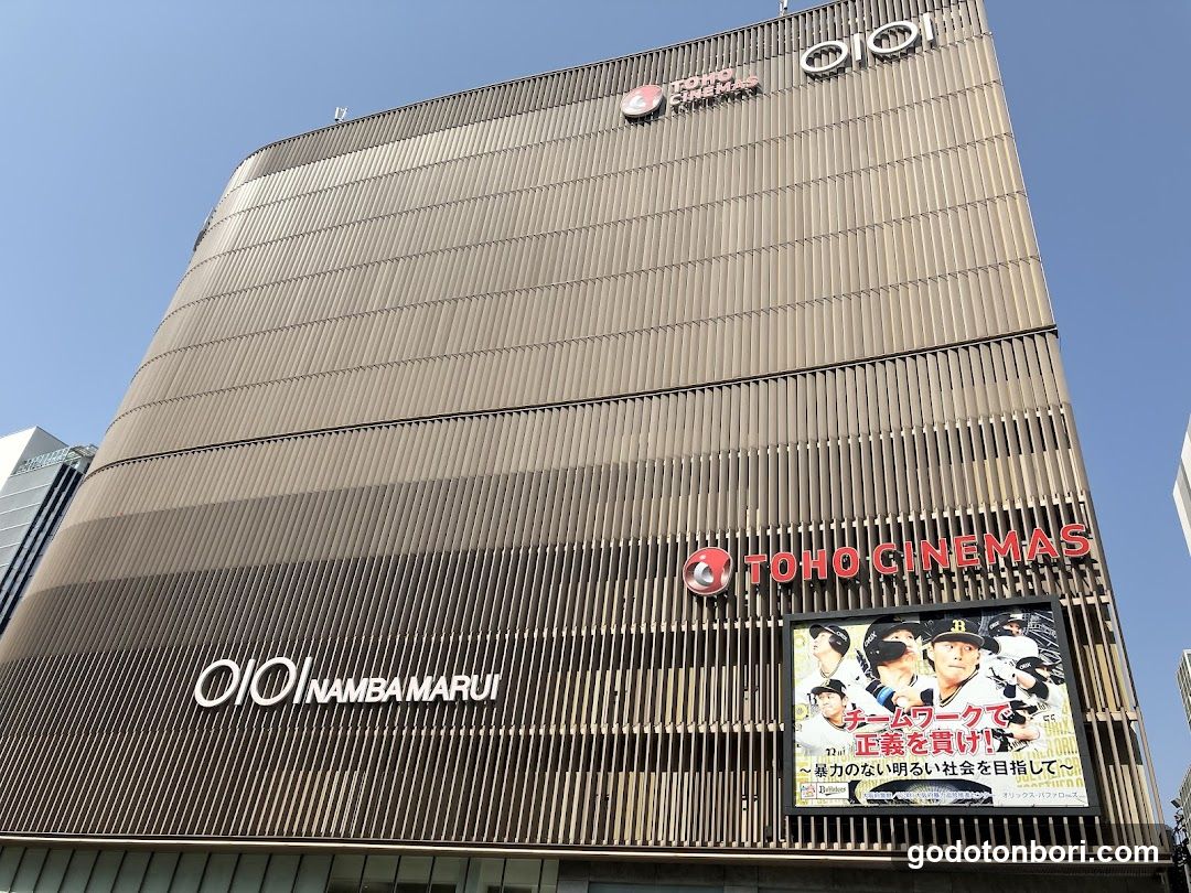 Low-angle shot of the facade of a multi-story building with horizontal slat cladding. Visible signage includes 'OIO NAMBAMARUI' and 'TOHO CINEMAS'. A large digital billboard displays an advertisement featuring baseball players in white uniforms, with Japanese text overlaying the image against a clear blue sky.