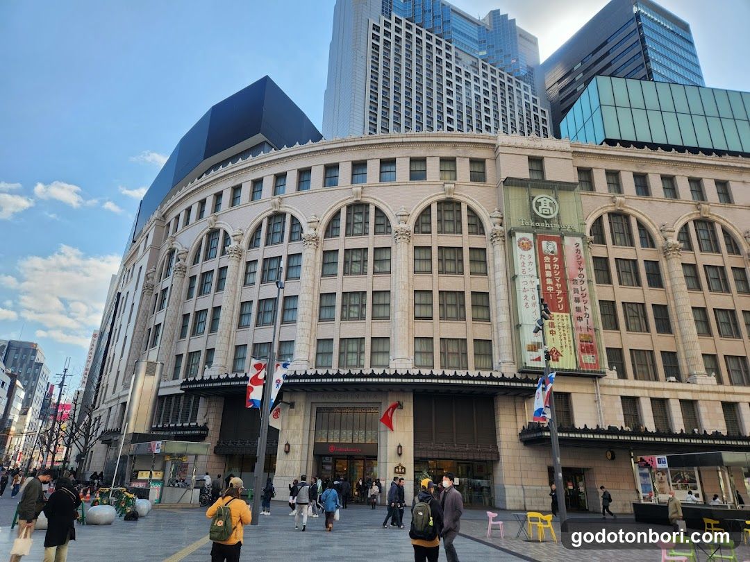 A wide-angle view of the Takashimaya department store entrance in Tokyo, Japan. The building features a curved, beige stone facade with arched windows and decorative columns. A large vertical banner displays Japanese text and English branding 'Takashimaya'. Several flags are visible near the entrance. Pedestrians walk on the paved plaza in front.