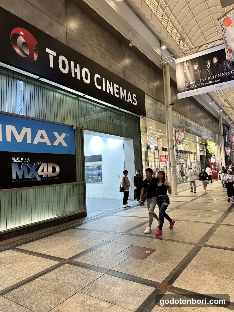Exterior view of TOHO CINEMAS storefront with visible signage including IMAX and MX4D logos. Pedestrians walking in front, illuminated entrance, and promotional banners hanging above.