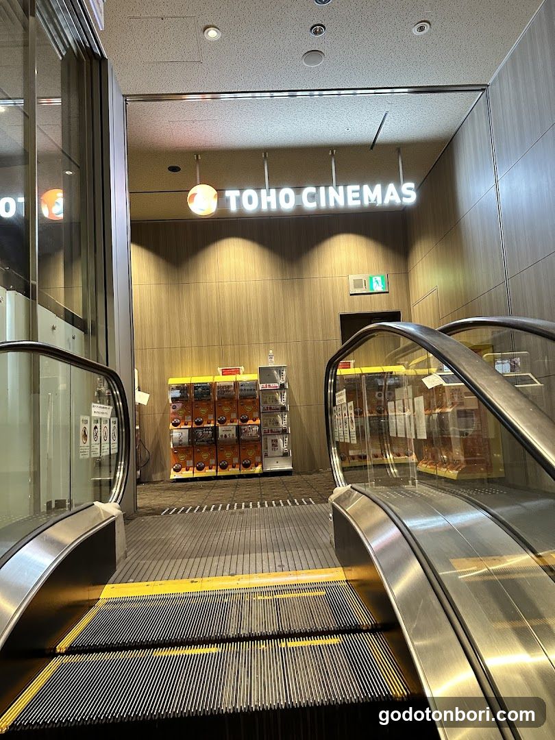 Photo of the entrance to a TOHO CINEMAS, showing escalators leading up to the cinema lobby with illuminated signage above and vending machines on either side.