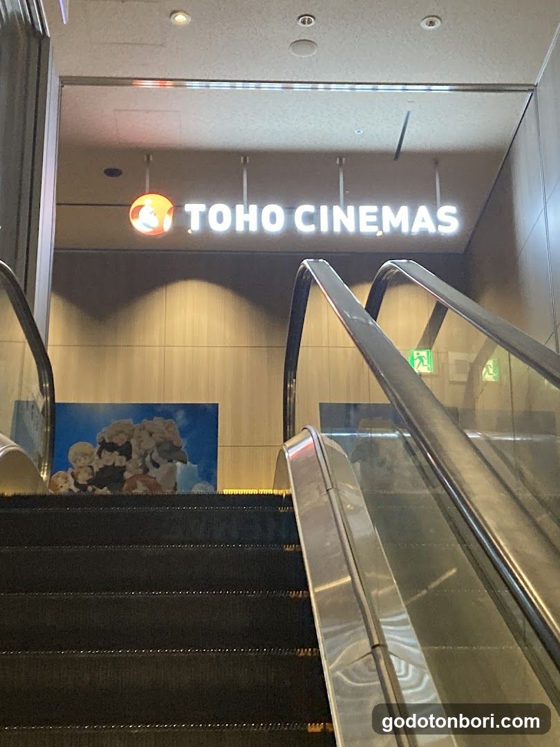 View of the TOHO CINEMAS entrance featuring a brightly lit sign with the orange logo above escalators leading to the cinema lobby. A promotional poster is visible on the wall beside the stairs.