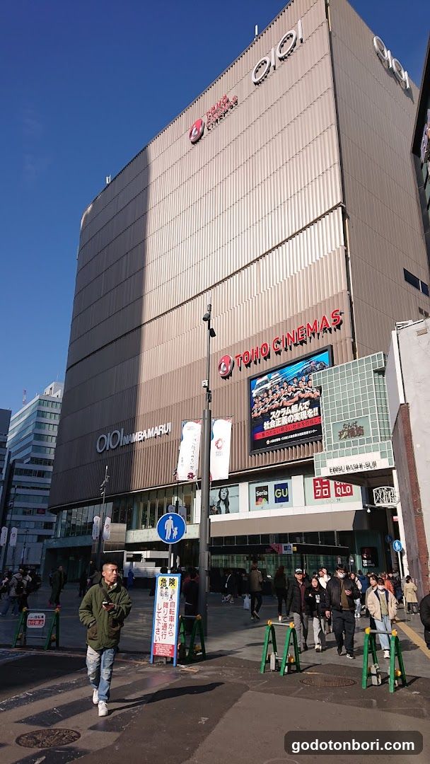 A tall, modern building with 'OIOI' and 'TOHO CINEMAS' signage prominently displayed on its facade. The ground floor features multiple shop entrances including GU, RLO, and other retail stores. A large digital screen displays movie advertisements. Pedestrians walk along the sidewalk in front of the entrance under a clear blue sky.