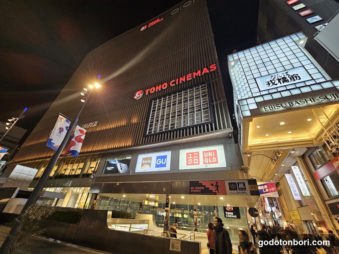 Night view of the Toho Cinemas building facade with illuminated signage, including 'TOHO CINEMAS' and adjacent 'EBISU BASHI-SUIJI'. Storefronts display Uniqlo and Seris branding. Pedestrians walk near entrance under warm lighting.