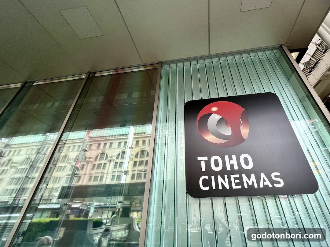 A low-angle shot of the TOHO CINEMAS storefront, featuring a modern glass facade with vertical slats and a prominent black sign displaying the red spherical logo and white lettering. Reflections of surrounding buildings are visible in the windows.