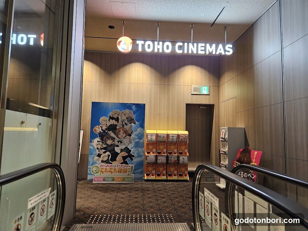 Entrance to TOHO CINEMAS with illuminated signage above the door, flanked by escalators. A large anime movie poster is displayed on the left wall, and stacks of merchandise boxes are arranged near the center. An exit sign is visible above a doorway in the background.