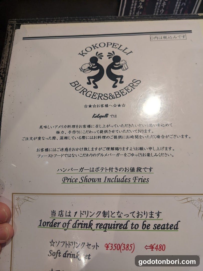 A close-up of a printed menu page from Kokopelli Burgers & Beers, featuring Japanese and English text. The top section displays the logo with two stylized figures holding drinks. Below are notices about tipping, service time, and price inclusion of fries. A section indicates one drink per order is required to be seated. At the bottom, a soft drink set is listed at ¥350 (385) + tax ¥480.