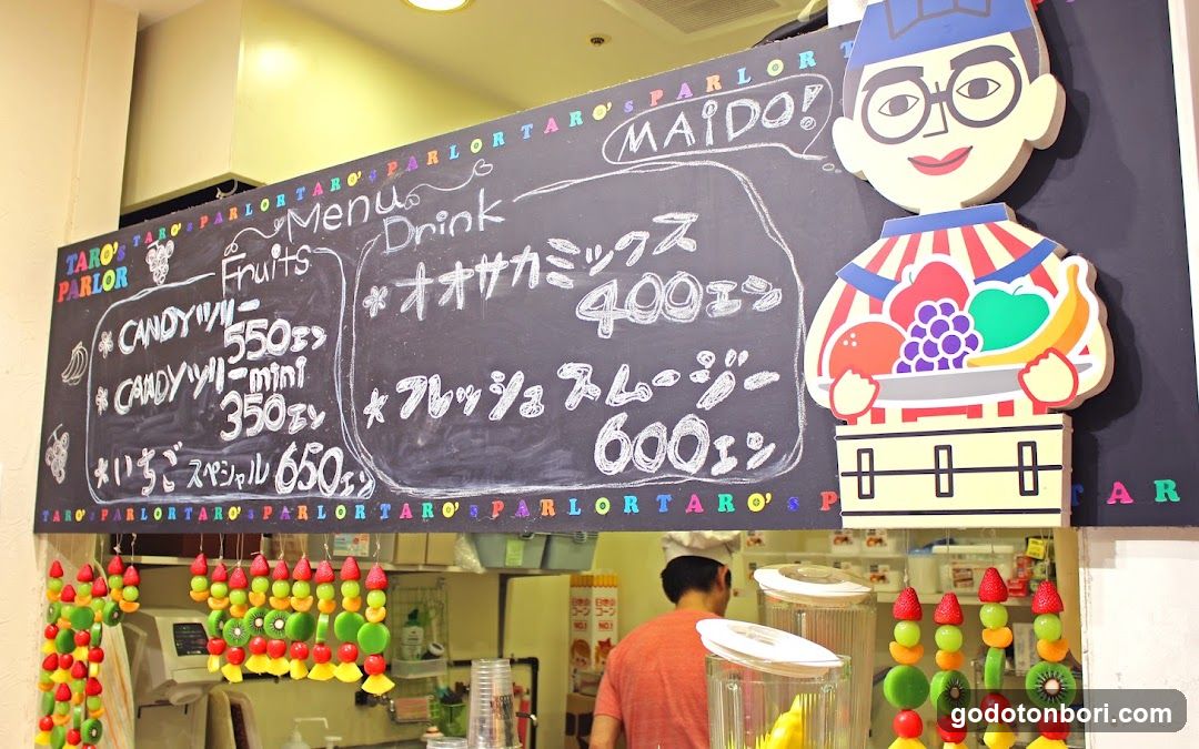 A chalkboard menu board displaying fruit and drink options with prices in Yen, featuring a cartoon character illustration on the right side.