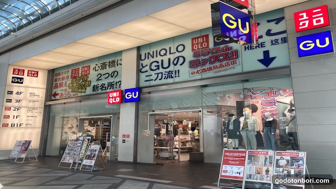 Exterior view of a shopping complex featuring UNIQLO and GU stores. Visible signage includes 'GU' in red and blue, 'UNIQLO' logo, and Japanese text promoting the collaboration between the two brands. Storefronts have glass entrances with mannequins displayed outside. Aftersales information boards are placed near the entrance.