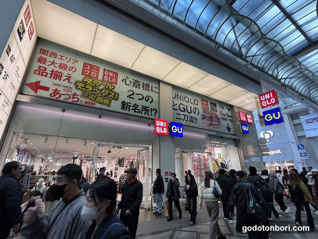 A busy street-level view of a Uniqlo and GU store entrance, featuring prominent signage with Japanese text advertising promotions like 'Kansai Area's largest product grab' and 'Two new spots in Kokubunji'. The storefront is bustling with shoppers, some wearing masks, under an arched glass ceiling structure.