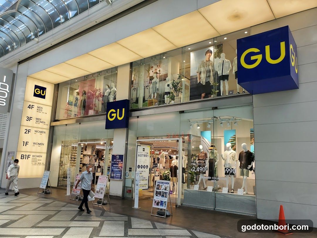 Exterior view of GU store with blue signage, glass facade, mannequins in window display, promotional posters inside, and directional signs indicating floors (1F-4F). A person walks past the entrance.