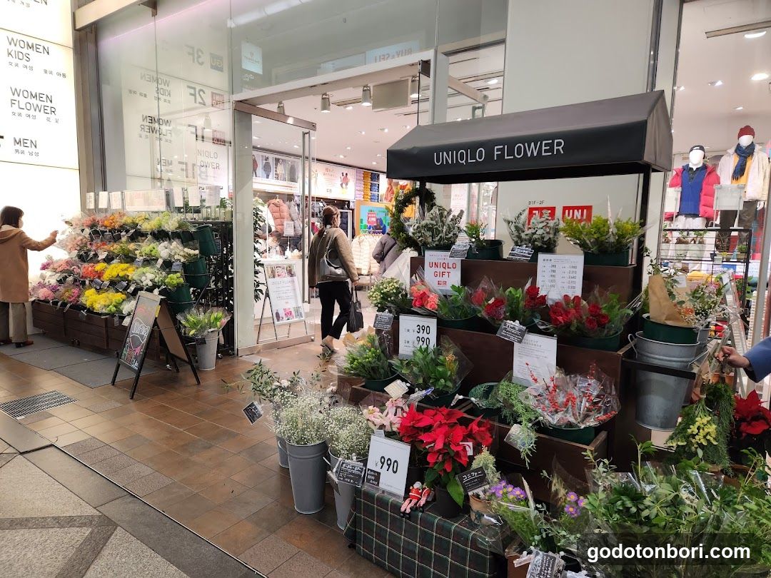 A flower shop display outside a store, featuring various floral arrangements and potted plants. A sign reading 'UNIQLO FLOWER' is visible above the display. Inside, customers browse through more flowers and merchandise. The storefront includes glass doors and windows displaying additional items.