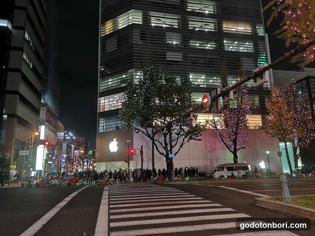 Night view of an Apple Store entrance with illuminated signage and storefront facade, surrounded by urban streetlights and pedestrians.