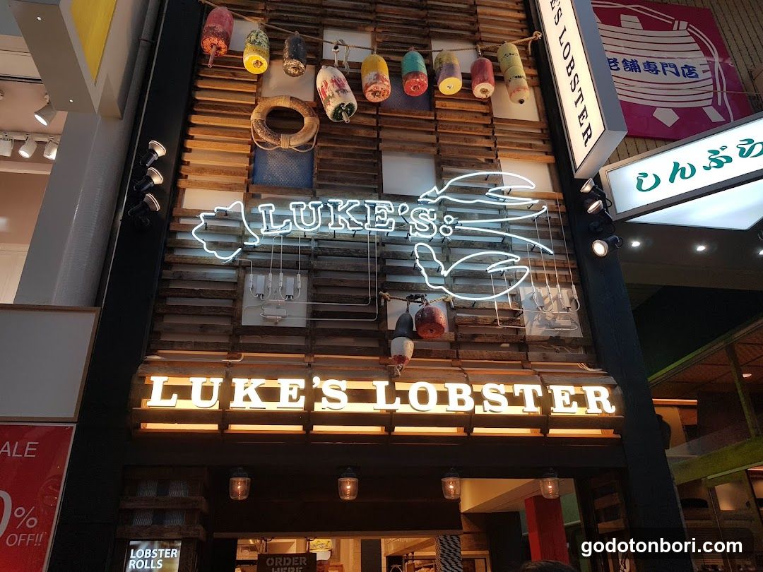 A storefront facade featuring a wooden slat wall with hanging colorful cylindrical decorations and neon outlines of lobsters. The sign reads 'LUKE'S LOBSTER' in illuminated white letters, with additional signage above indicating Japanese characters. Small lanterns hang near the entrance.