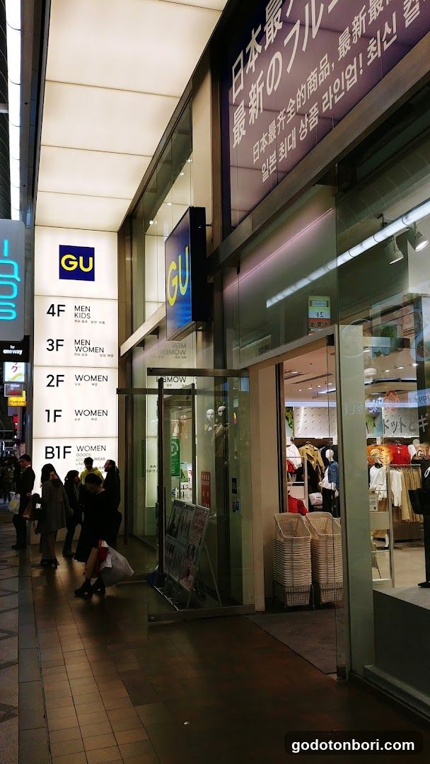 Exterior view of a GU store entrance with visible signage indicating floors (B1F, 1F, 2F, 3F, 4F) and sections for men, women, and kids. The storefront features glass doors and windows displaying clothing items inside. A stack of white baskets is placed near the entrance.
