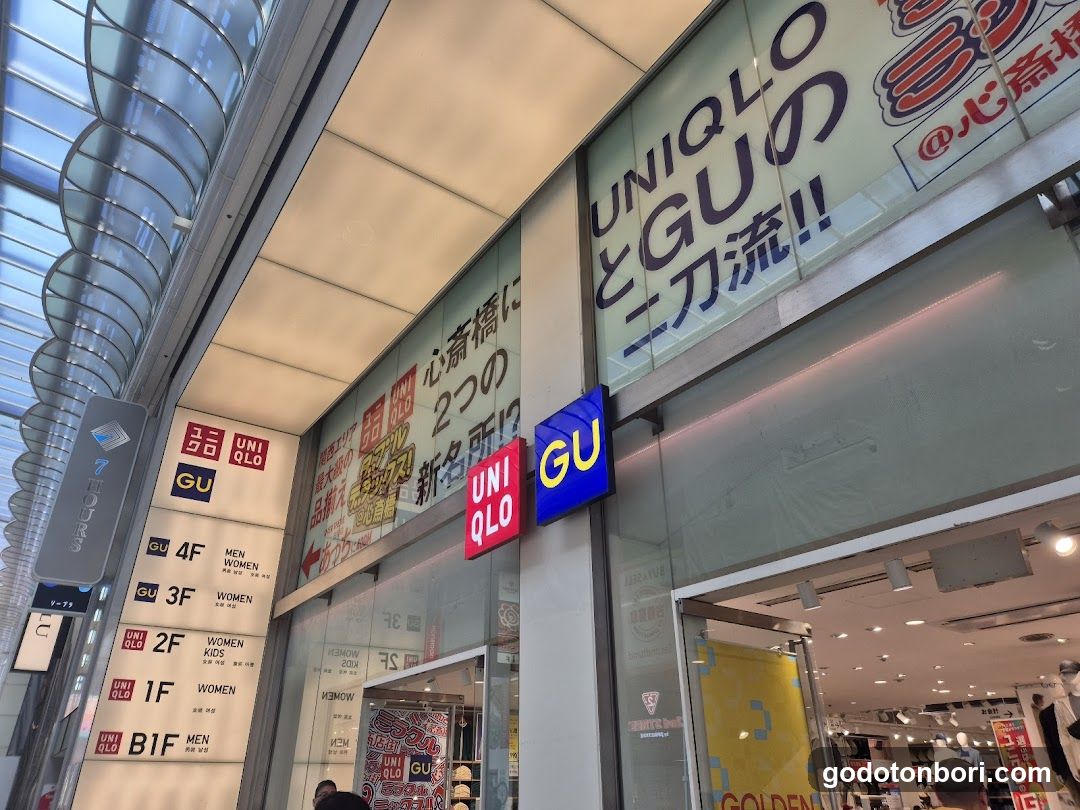 The image shows the exterior of a Uniqlo store, featuring signage with 'UNIQLO' and 'GU' logos prominently displayed. The storefront includes directional signs indicating different floors (B1F to 4F) for men's and women's sections. A large banner advertises 'UNIQLO と GU の二刀流!!', suggesting a collaboration or dual-brand presence. The entrance is visible with glass doors leading into the brightly lit interior.