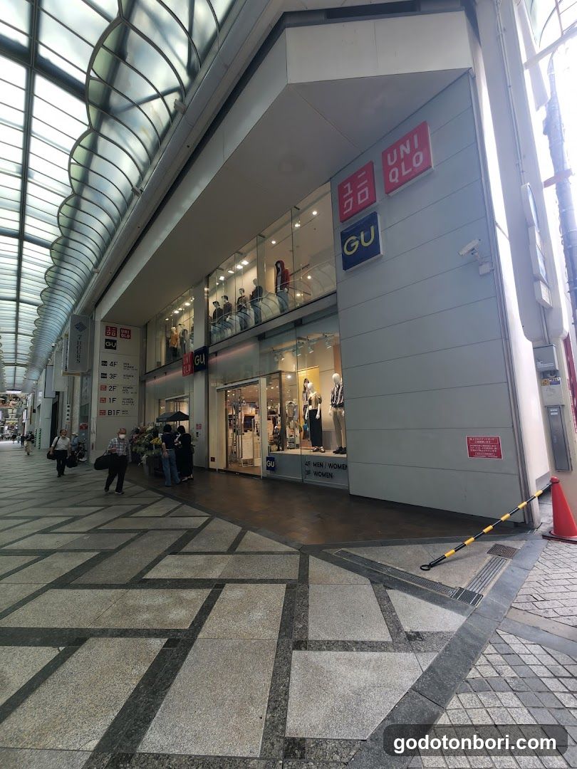 The image shows the exterior of a Uniqlo store with adjacent GU branding visible on the building facade. The storefront features large glass windows displaying mannequins inside, with clear signage above the entrance. A few pedestrians are walking nearby under an arched walkway structure.