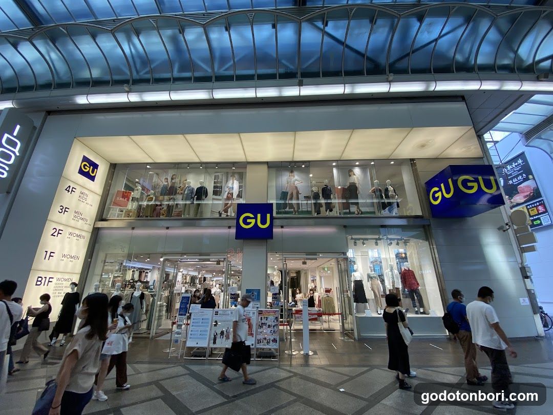 A modern two-story clothing store with large glass windows and doors, featuring GU branding on blue signs above the entrance and on the facade. The interior is visible through the glass, showing mannequins dressed in various outfits. People are walking by outside, and there are promotional posters near the entrance.