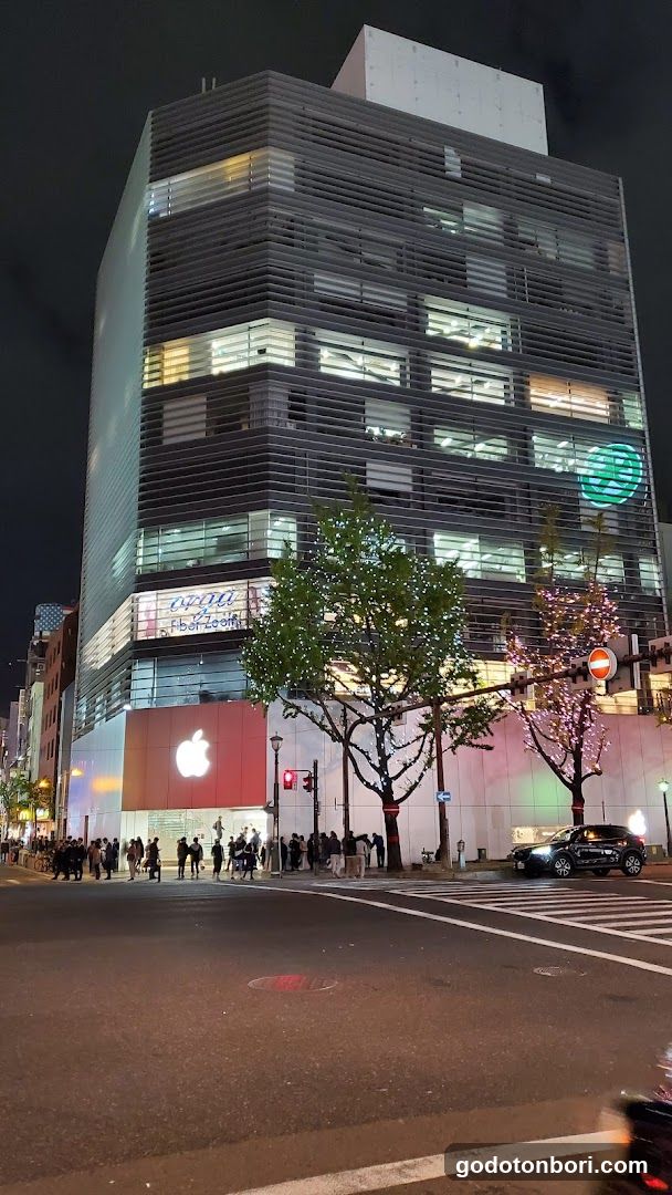 A modern multi-story building with a prominent Apple logo on the ground floor facade, illuminated by artificial lighting against a dark night sky. The upper floors feature horizontal slats and lit windows. A crowd of people is gathered near the entrance, and decorative lights adorn nearby trees.