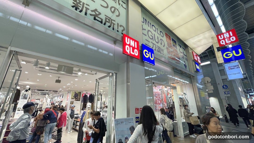 The image shows the exterior of a Uniqlo and GU store, with visible signage on the building facade. The entrance is open, revealing the interior with clothing racks and mannequins. People are walking by or entering the store.
